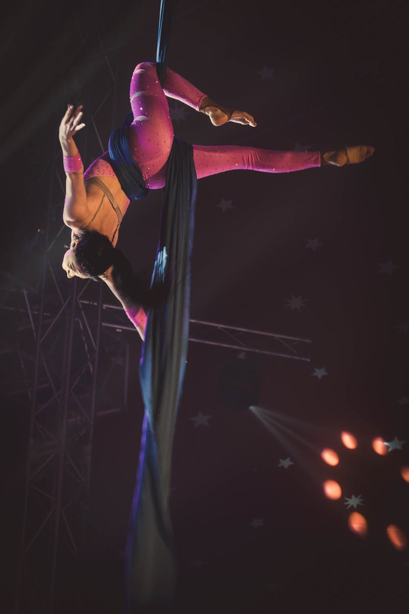 A skilled acrobat performs upside down on silk ribbons under dramatic lighting.