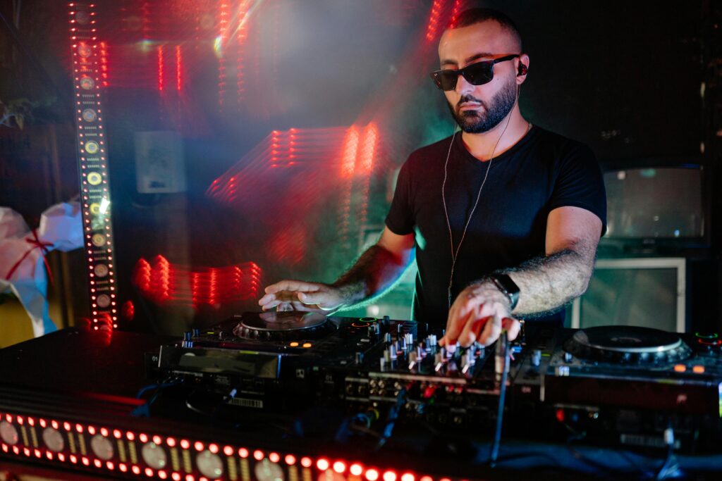 A DJ in sunglasses and black shirt playing electronic music at a vibrant nightclub.